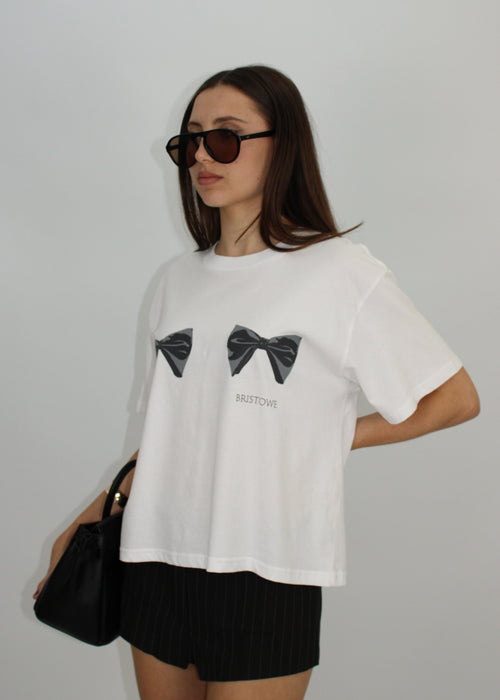 BRISTOWE Beau Tee in white — three-quarter front view, oversized white tee with black double bow graphic and BRISTOWE logo, styled with black shorts and tortoiseshell sunglasses